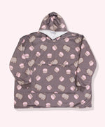 Pusheen Wearable Blanket Hoodie lies on a light-pink surface. The oversized pullover has a hood, oversized sleeves, a large front pocket, and an all-over-print Pusheen the Cat with pink macarons. 