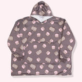 Pusheen Wearable Blanket Hoodie lies on a light-pink surface. The oversized pullover has a hood, oversized sleeves, a large front pocket, and an all-over-print Pusheen the Cat with pink macarons. 