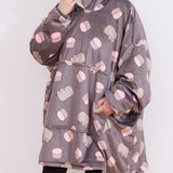 Model wears a Macaron Blanket Hoodie. With the sleeves scrunched up, the model holds the side of hood with one hand and has her other hand in the large front pocket. The light grey onesie has an all over print of Pusheen the Cat and pink macarons. The hoodie is oversized and lined with sherpa for a cozy fit. 