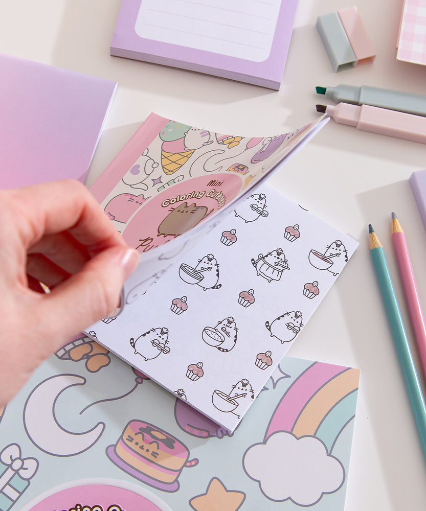 Model holds open the Pusheen activity book showcasing one of the coloring pages. The open page shows a black outline Pusheen carrying cupcakes, mixing batter, and mini cupcakes.