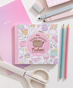 Mini coloring book with Pusheen and character illustrations on a white desktop with stationery items including pencils, scissors, and markers.