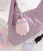 Plush keychain attached to a purple bag with a notebook and sunglasses on a light surface.