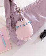 Plush keychain attached to a purple bag with a notebook and sunglasses on a white surface.