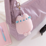 Plush keychain attached to a purple bag with a notebook and sunglasses on a white surface.