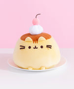 Pudeen Squisheen sits on a cream chair to show the size of the plush. The light-yellow pudding-shaped plush has a light caramel top with dark brown embroidery features for Pusheen's eyes, mouth, and whiskers. 