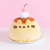 Front view of Pusheen Pudeen Squisheen. Pusheen the Cat takes the form of a pudding dessert. The dessert-inspired plush has light brown embroidery to show off Pusheen’s classic head stripes while her nose, mouth, eyes, and whiskers are in a dark brown embroidery thread color. The yellow plush sits atop a white plate. 