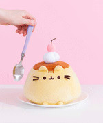 Gif of Pusheen Pudeen Squisheen being tapped with a silver and purple metal spoon held in a model’s hand. Pusheen the Cat takes the form of a flan pudding dessert treat. The top of the plush is a warm brown color to mimic a caramel top while the light-yellow bottom represents the pudding. The plush is topped with a whipped cream dollop and cherry plush.