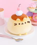 Pudeen Squisheen plush sits atop a white plate surrounded by a silver and pink spoon, pink storage cannister, and cupcakes and donuts. The Squisheen base is light yellow with a warm golden brown to mimic caramel topping. A white whipped cream dollop and cherry plushes sit atop the pudding-inspired plush.  