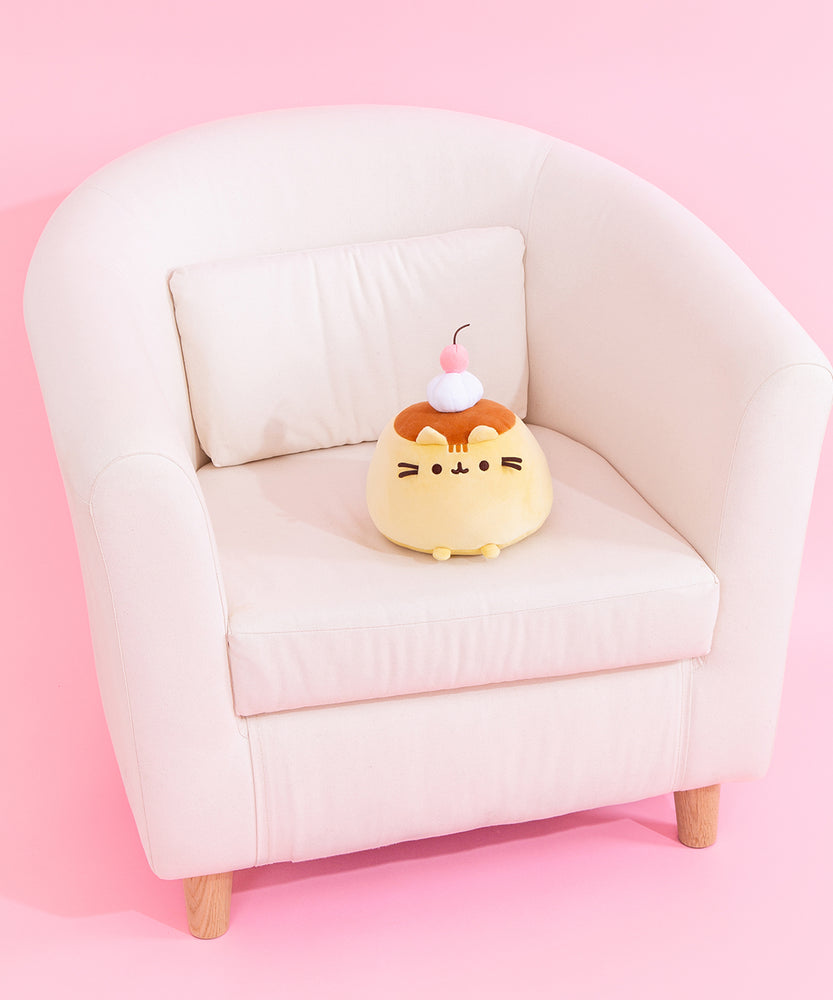 Pudeen Squisheen sits on a cream chair to show the size of the plush. The light-yellow pudding-shaped plush has a light caramel top with dark brown embroidery features for Pusheen's eyes, mouth, and whiskers. 