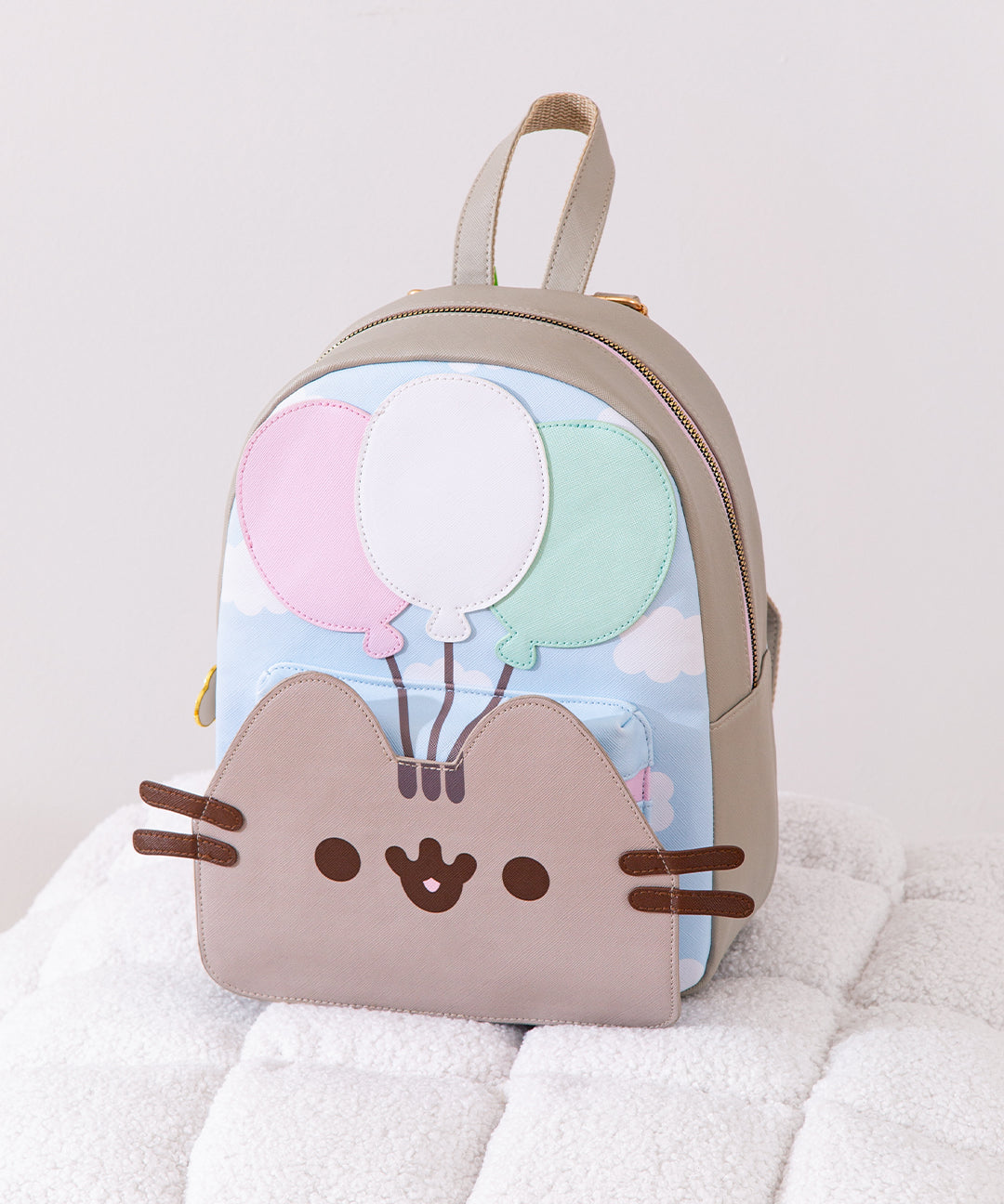Pusheen 15th Anniversary Balloons Backpack sits on a white poof. The gray backpack has Pusheen the Cat's face on the front of the pack paired with three balloons.