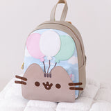 Pusheen 15th Anniversary Balloons Backpack sits on a white poof. The gray backpack has Pusheen the Cat's face on the front of the pack paired with three balloons.