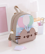 The Pusheen 15th Anniversary Balloons Backpack sits on a white desktop surrounded by office supplies. The front of the backpack has a raised portion showcasing a light pink, white, and mint green balloons trio. 