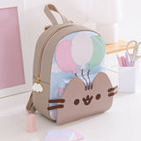 The Pusheen 15th Anniversary Balloons Backpack sits on a white desktop surrounded by office supplies. The front of the backpack has a raised portion showcasing a light pink, white, and mint green balloons trio. 