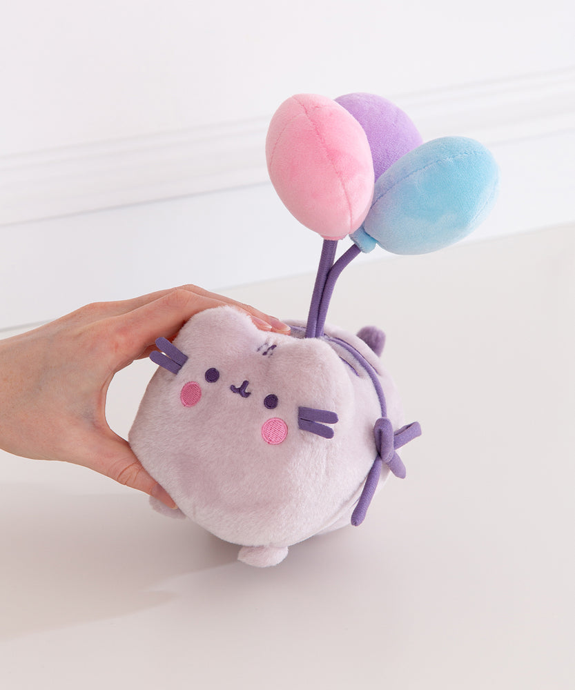 Pusheen plush toy with balloons held by a hand on a white background