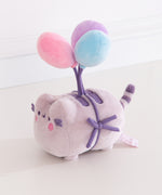 Quarter side view of the Pusheen 15th Anniversary Balloons Plush toy. Pusheen the cat is tied to three multi-colored balloons. Her normally brown details are dark purple.