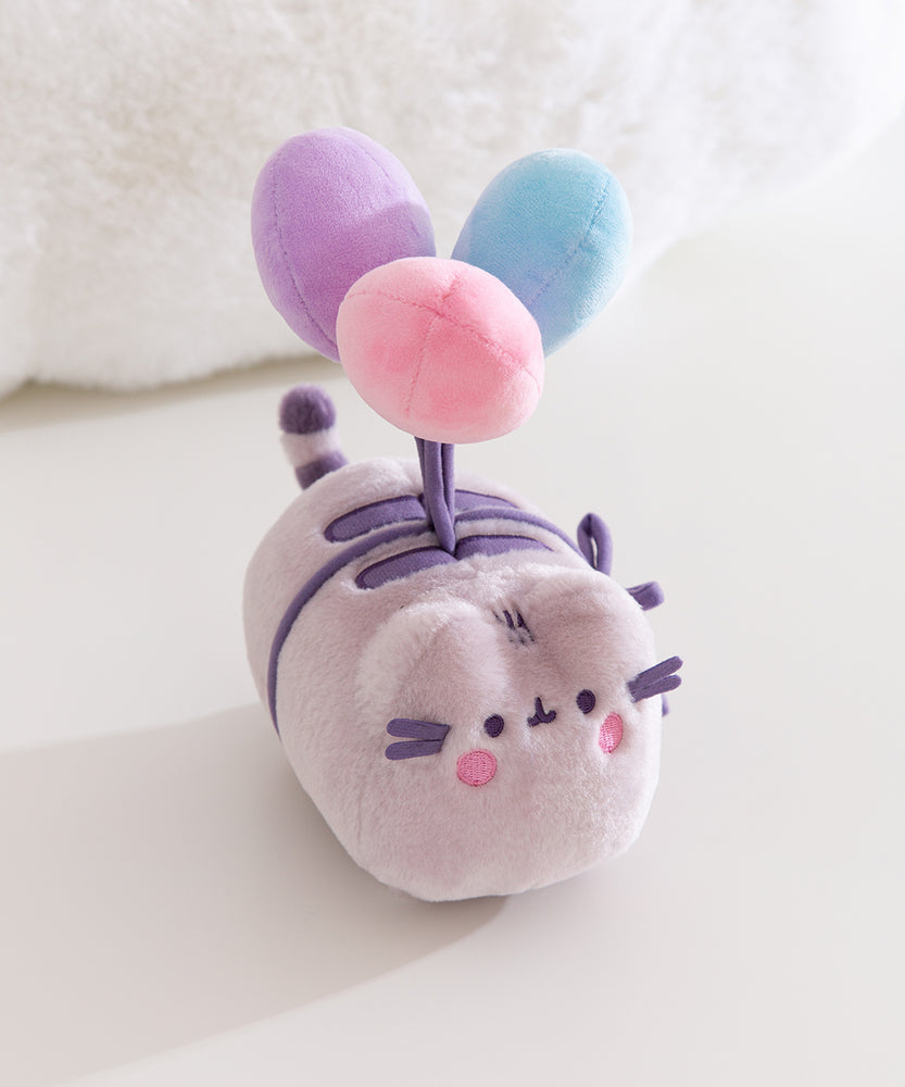 Aerial view of the plush toy showing Pusheen the Cat blushing while attached to blue, purple, and pink plush balloons. 