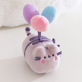 Aerial view of the plush toy showing Pusheen the Cat blushing while attached to blue, purple, and pink plush balloons. 