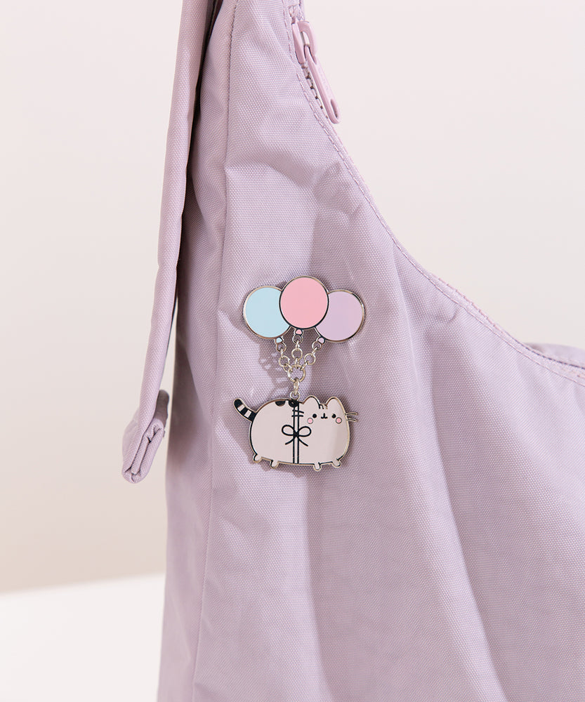 A blushing Pusheen the Cat hangs from three pastel blue, pink, and purple balloons and the pin is attached on a light purple bag.