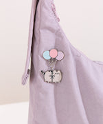 A blushing Pusheen the Cat hangs from three pastel blue, pink, and purple balloons and the pin is attached on a light purple bag.