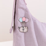 A blushing Pusheen the Cat hangs from three pastel blue, pink, and purple balloons and the pin is attached on a light purple bag.
