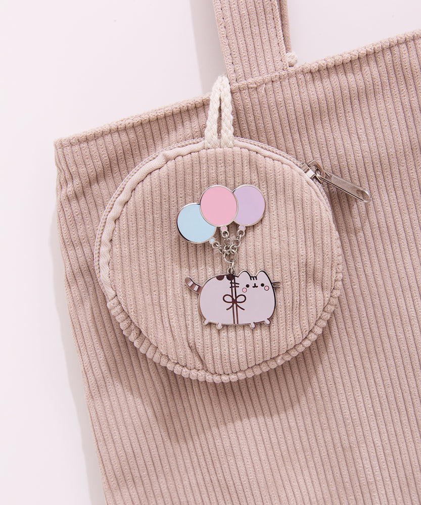 Pin of Pusheen dangling from three balloons. The silver outlined pin is attached to a corduroy bag.