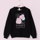 The Pusheen 15th Anniversary Tee hangs on a light pink hanger. The black sweatshirt features a large graphic of Pusheen with a trio of balloons and surrounded by fireworks.