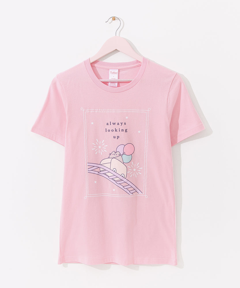 The Pusheen 15th Anniversary Tee hangs on a light pink hanger. The pink tee features a large graphic of Pusheen riding a rollercoaster, holding balloons, and surrounded by stars and fireworks.