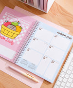 Open planner to March 2026. The fruit themed calendar page is paired with a 7-day calendar view. The planner lies on a wooden desk next to a pen and keyboard.
