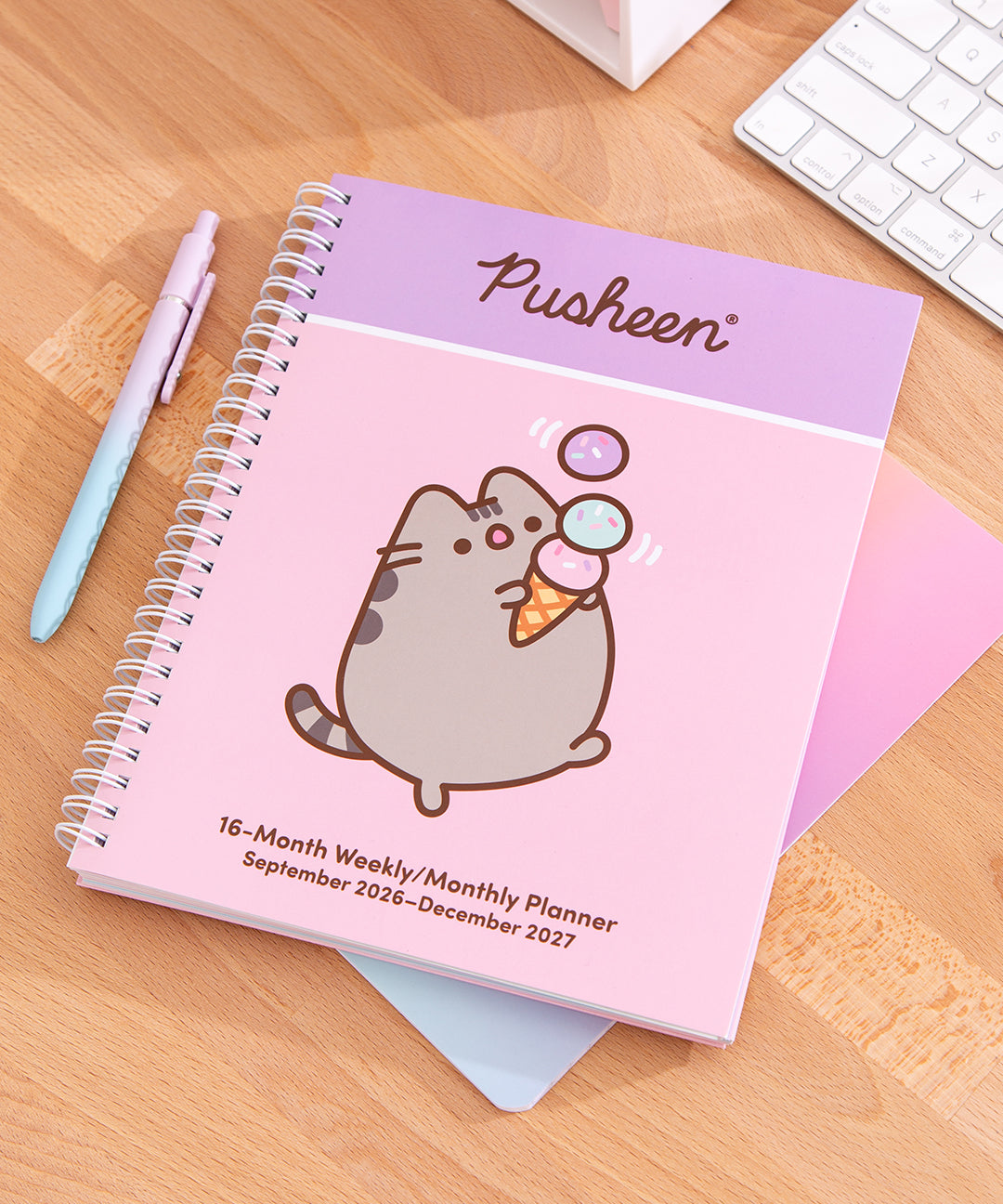 Front cover of the Pusheen 16-month planner on a wooden desk with stationery items. The pink planner shows a graphic of Pusheen carrying a three-scoop ice cream cone.