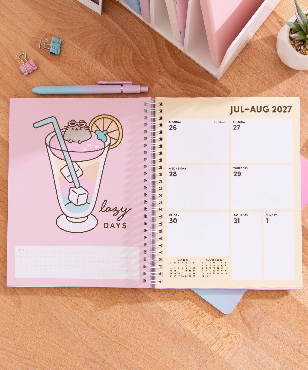 Calendar open to the last week of July 2027. The page spread is tropical vacation themed and shows Pusheen in an ombre drink glass.
