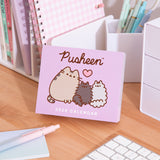 Pusheen 2026 daily calendar box on a desk with stationery items. The front graphic on the purple box shows Pusheen the Cat with her siblings Pip and Stormy. 