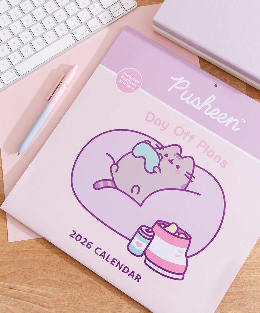 Front cover of the Pusheen 2026 wall calendar on a desk with a keyboard and pen. The front cover includes a graphic of a cat holding a gaming controller and sitting in a large bean bag chair surrounded by snacks. 