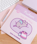 Front cover of the Pusheen 2026 wall calendar on a desk with a keyboard and pen. The front cover includes a graphic of a cat holding a gaming controller and sitting in a large bean bag chair surrounded by snacks. 