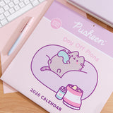 Front cover of the Pusheen 2026 wall calendar on a desk with a keyboard and pen. The front cover includes a graphic of a cat holding a gaming controller and sitting in a large bean bag chair surrounded by snacks. 