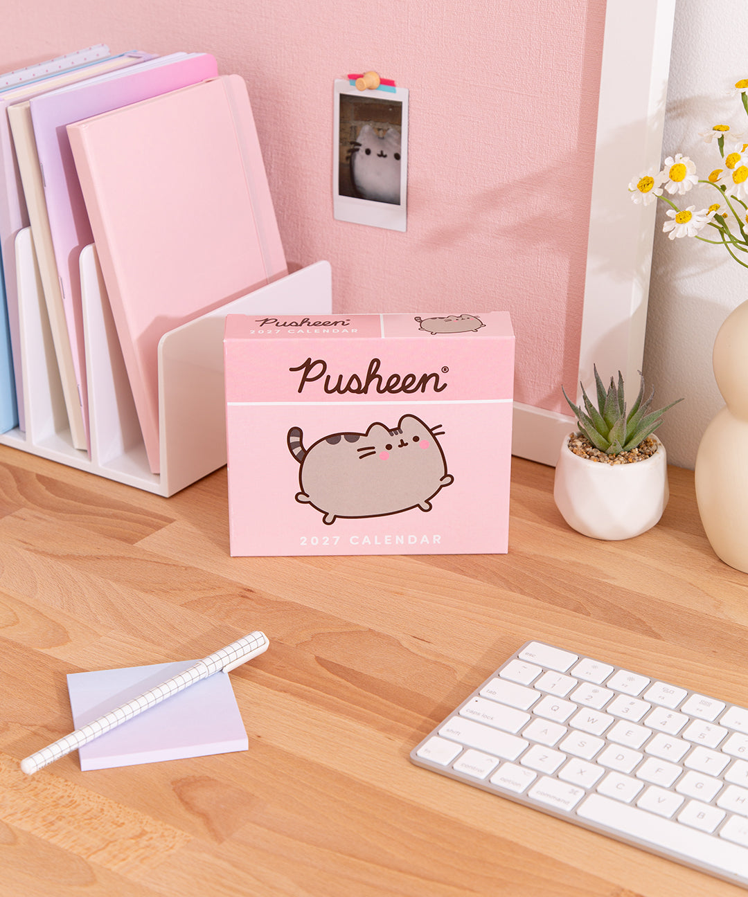 Pusheen 2027 daily calendar box on a desk with stationery items. The front graphic on the pink box shows Pusheen the Cat leaping and blusing. 