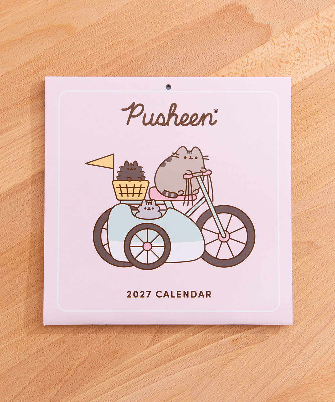 Front cover of the Pusheen 2027 mini wall calendar on a desk. The front cover includes a graphic of Pusheen riding a bicycle with Pip and Stormy in tow. The front also includes a Pusheen logo and text saying "2027 calendar".