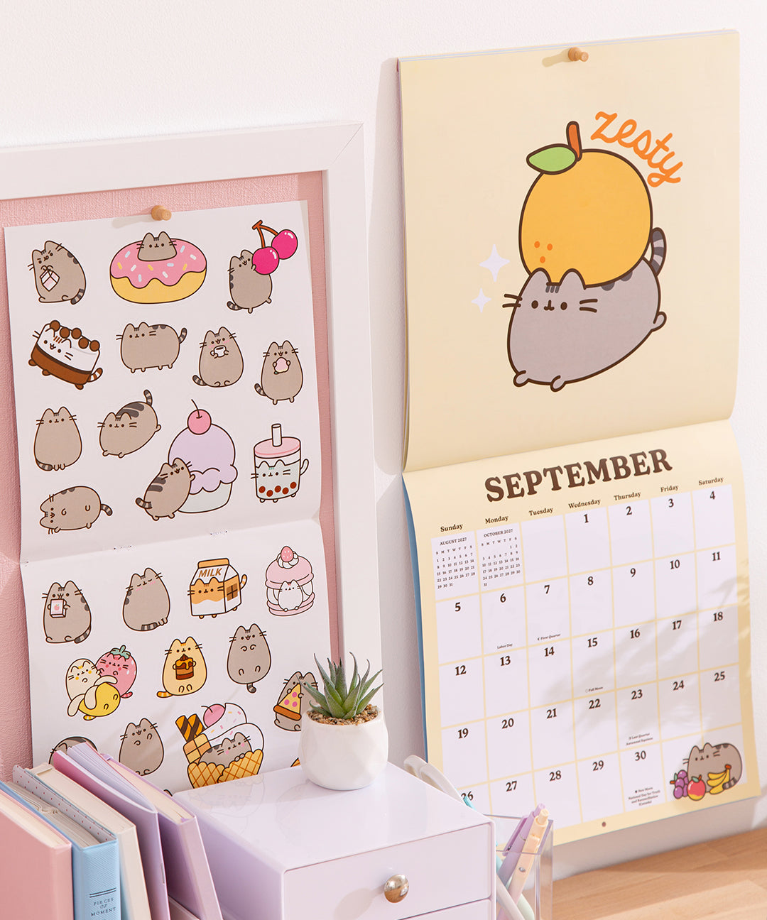 2027 Pusheen Calendar open to September 2027 hangs on a wall. Next to the wall calendar is the included sticker sheet.