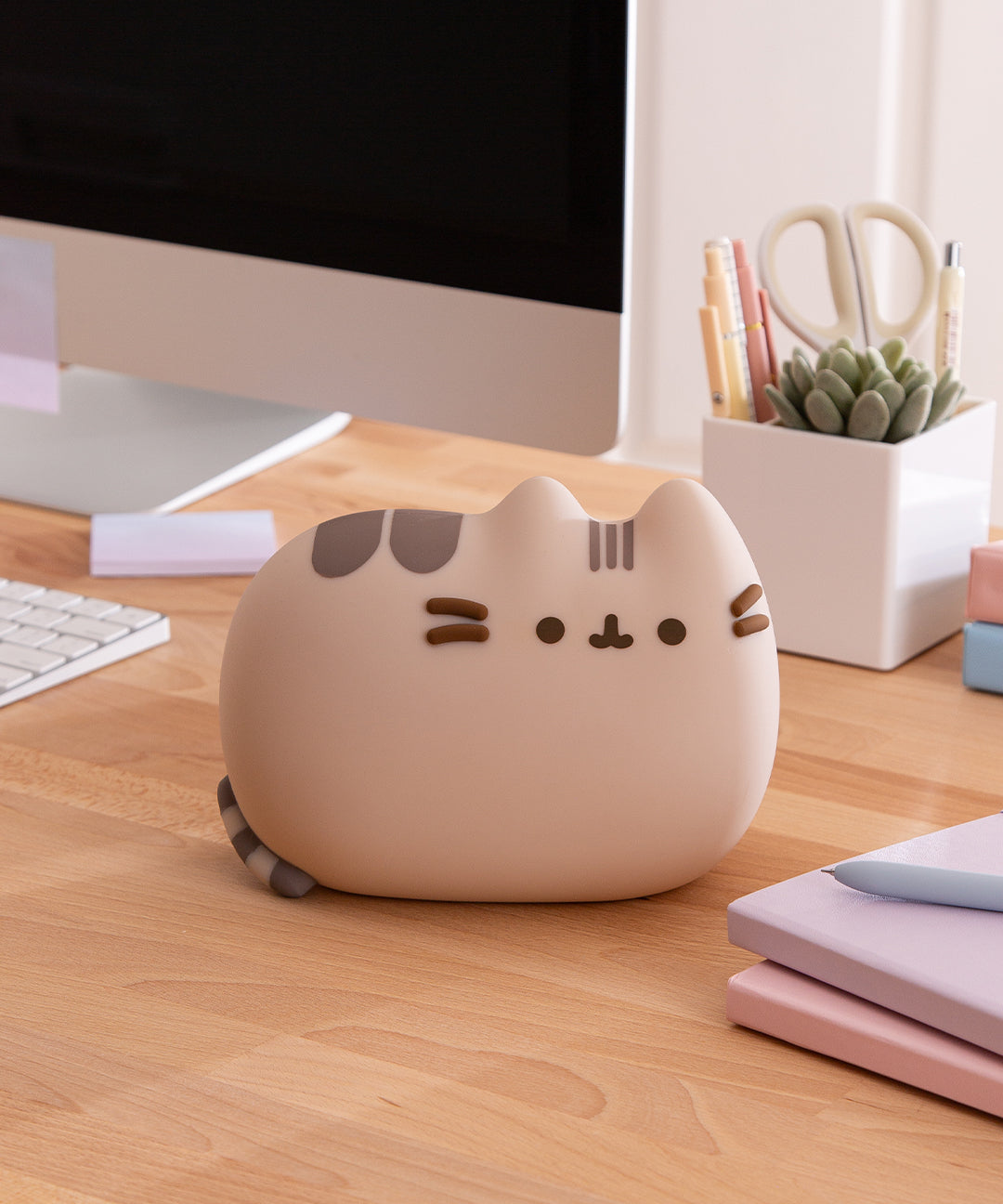 Pusheen Lamp sits on a desktop. The grey and brown tabby cat is laying down with her striped tail at her back.