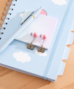 Pair of earrings with Pusheen the Cat dangling from pink balloons. The studs are angled to the side on a notebook with cloud designs.