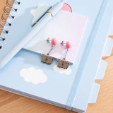 Pair of earrings with Pusheen the Cat dangling from pink balloons. The studs are angled to the side on a notebook with cloud designs.