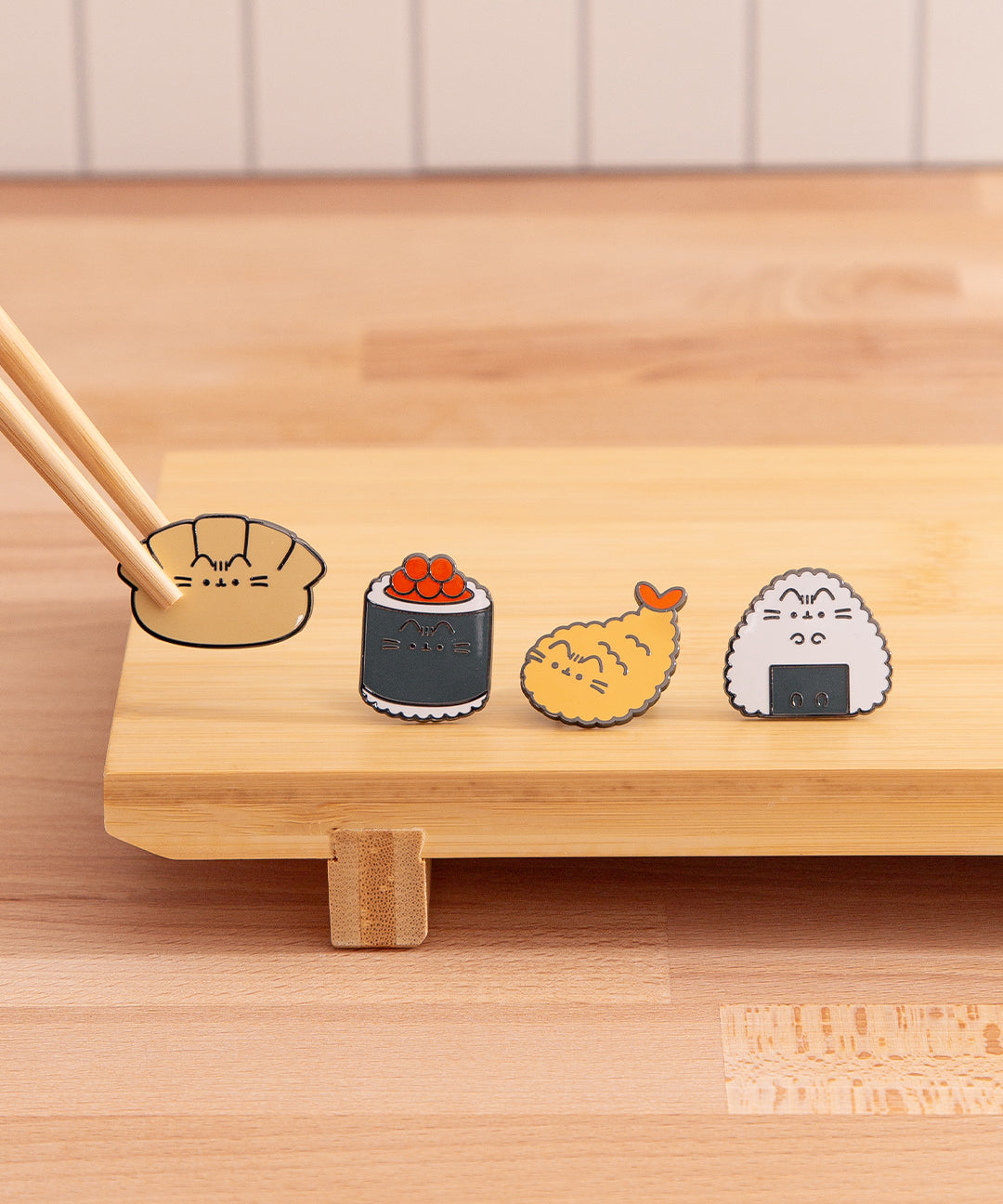 Close-up view of the Pusheen bento box pins on a kitchen top. A pair of chopsticks holds the gyoza dumpling pin to show the size of the pins.