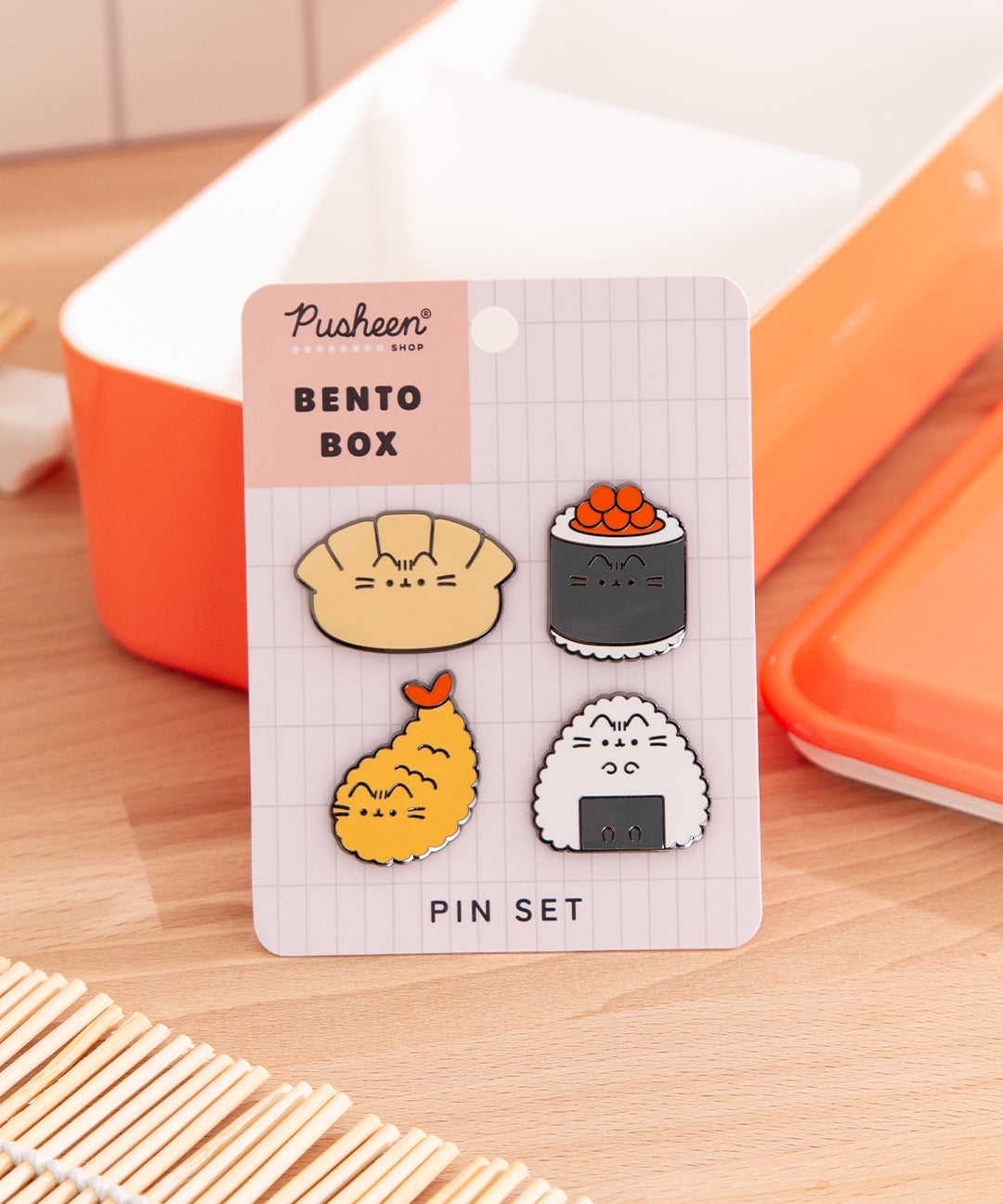 Pusheen Bento Box Pin Set is propped up by an orange lunch box. The four Bento Box Pusheen pins are attached to a backer card with the product name in dark brown color. The bento box items included in this pin set are gyoza, ikura sushi, shrimp tempura, and onigiri.