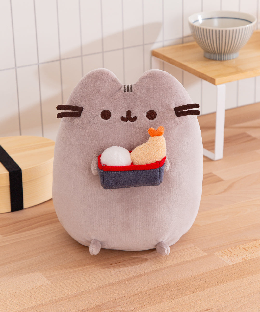 Front view of the Pusheen Bento Box Plush where a gray tabby cat holds a bento box including a rice ball and shrimp tempura.
