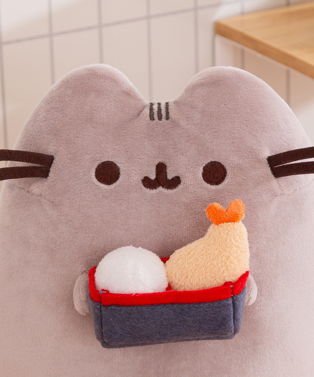 Close-up of the Pusheen Bento Plush. The cat is holding a gray and red bento box that contains a rice bal and shrimp tempura.