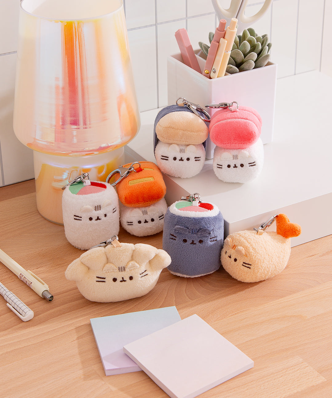 Front view of Bento Box Pusheen Surprise Plush assortment. Plush keychains sit on multi-level white pedestal in front of a desktop background. Keychains feature various bento box items including tamago sushi, tuna sushi, salmon sushi, uramaki sushi, maki sushi, gyoza, sushi roll, and shrimp tempura.