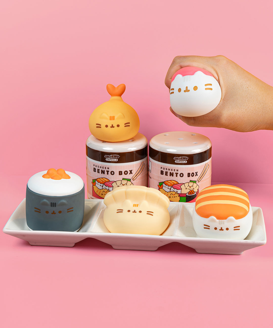 Front view of all Bento Box Pusheen Surprise Squishy including the blind box capsule. The squishies include a salmon sushi, tuna sushi, shrimp tempura, gyoza and ikura sushi.