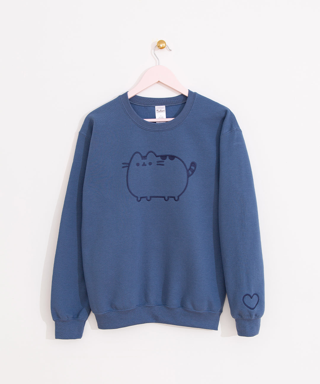 Pusheen Embroidered Unisex Sweatshirt hangs on a pink hanger in front of a white wall. The indigo blue sweatshirt features Pusheen embroidered in a dark blue thread on the front of the sweatshirt with a heart outline embroidered on the wearer’s left sleeve near the cuff. 