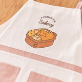 Pusheen Apron lies on a wooden tabletop. Printed in the graphic on the chest of thw apron is Pusheen the Cat taking the form of a baguette, loaf of bread, and croissant sitting in a brown bread basket. 