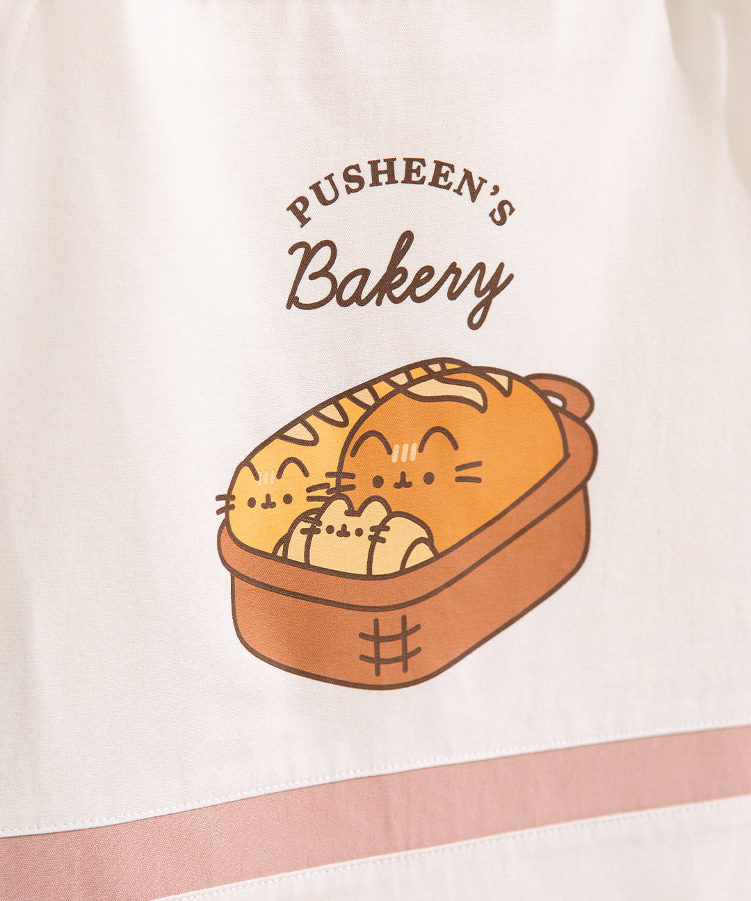 Close-up of the graphic printed on the chest of the apron. The graphic says Pusheen's Bakery and features a brown bread basket with three bread items: baguette, loaf, and croissant. 