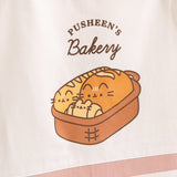 Close-up of the graphic printed on the chest of the apron. The graphic says Pusheen's Bakery and features a brown bread basket with three bread items: baguette, loaf, and croissant. 